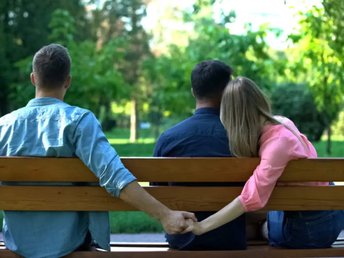 Pierwsze oznaki zdrady żony: kobieta siedzi na ławce z innym mężczyzną, trzymając go za rękę, podczas gdy jej mąż siedzi obok, nieświadomy.