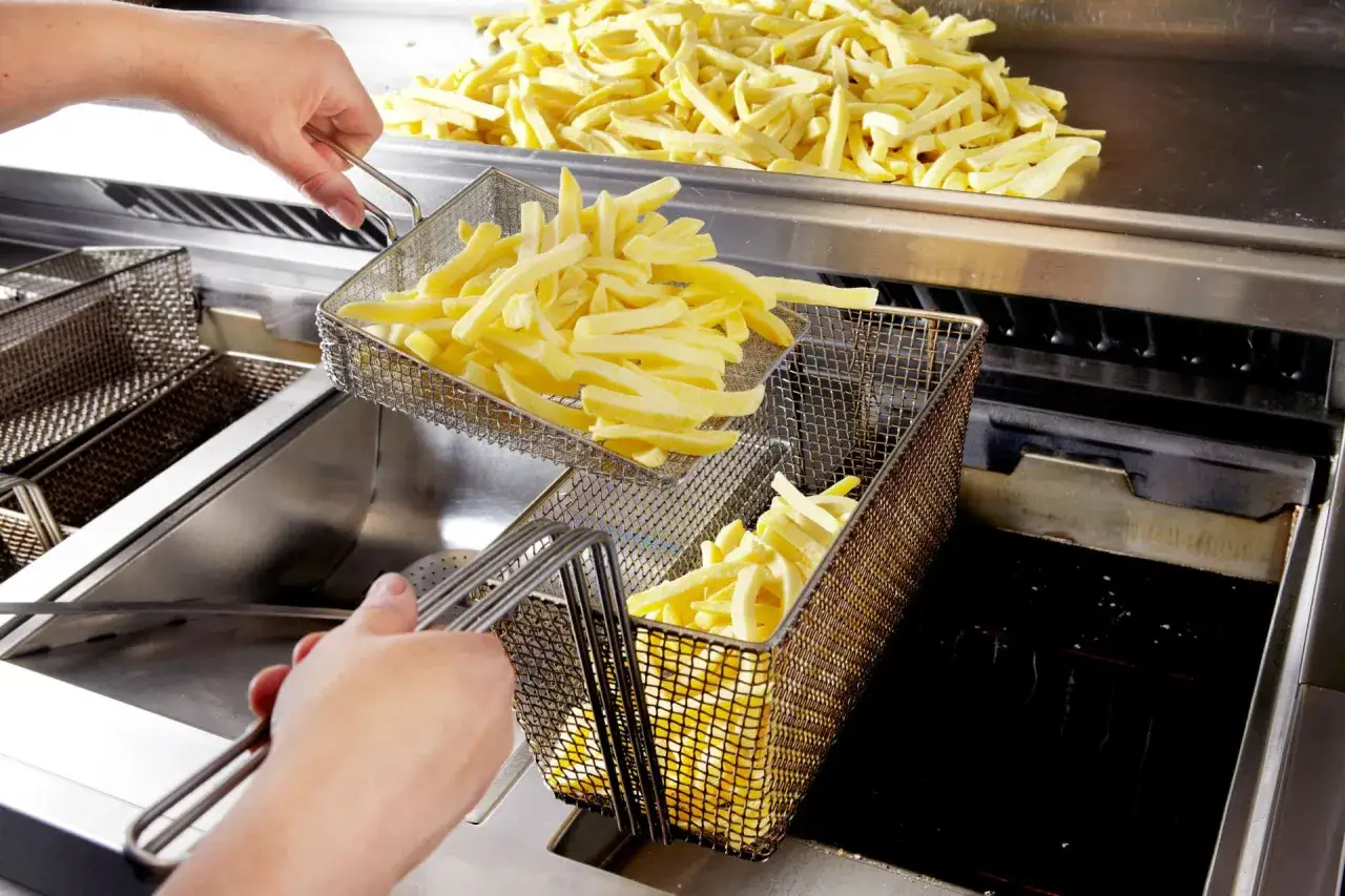 Frytki w koszyku do smażenia, a obok góra surowych ziemniaków. Widać też gorący olej, który może się pienić, gdy wrzucamy do niego ziemniaki.