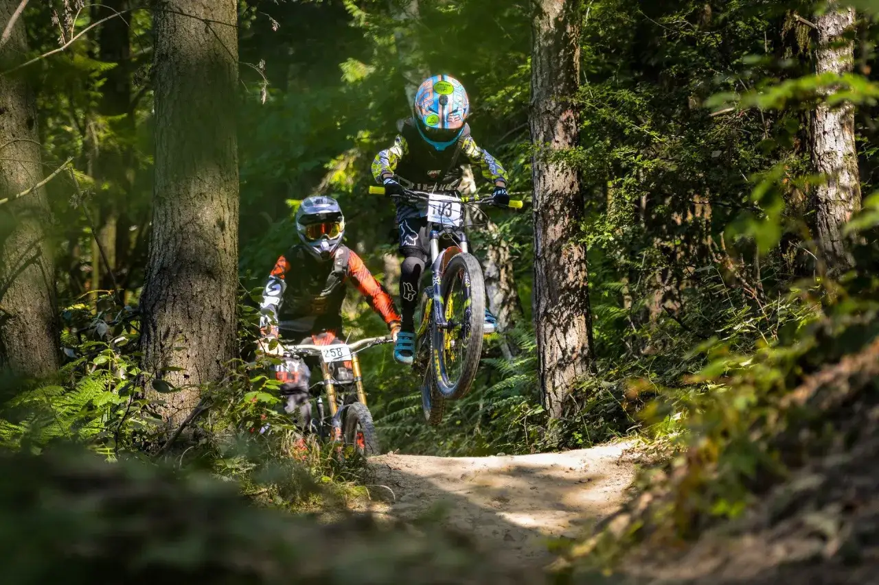 Dwóch rowerzystów w kaskach i ochraniaczach pokonuje leśny zjazd w bike parku Kasina. Jeden z nich jest w powietrzu.