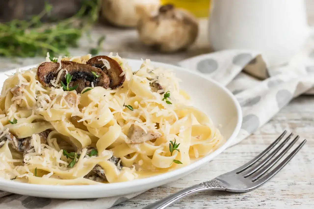 Tagliatelle z pieczarkami i kurczakiem, posypane parmezanem i świeżymi ziołami, prezentują się apetycznie na białym talerzu.
