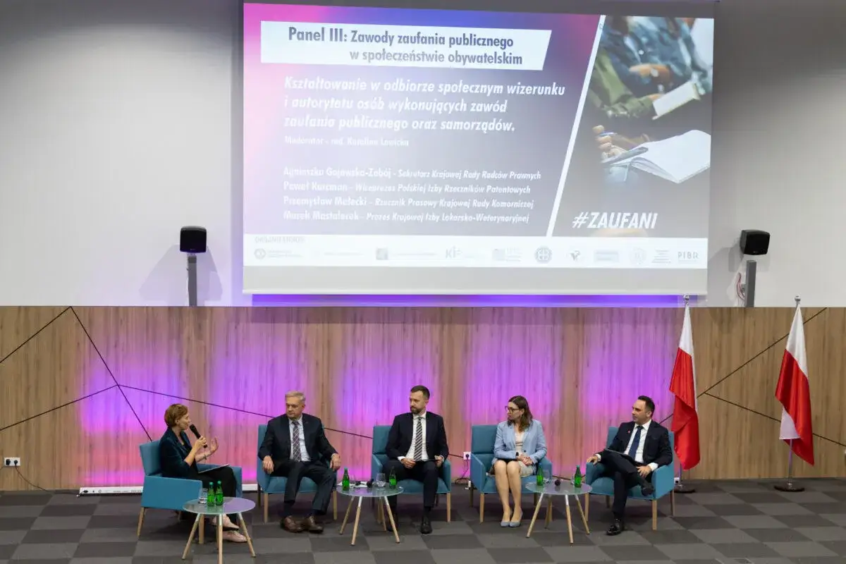 Panel dyskusyjny o kształtowaniu wizerunku osób zaufania publicznego. Uczestnicy siedzą na kanapach, na ekranie hasło #ZAUFANI.