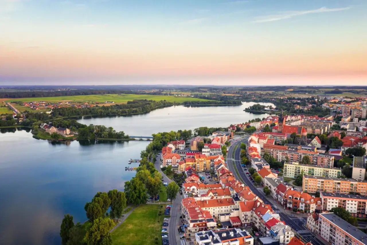 Widok na miasto nad jeziorem w ciepły wieczór. Dachy kamienic i zieleń otaczają wodę, tworząc malowniczy krajobraz Warmii i Mazur.