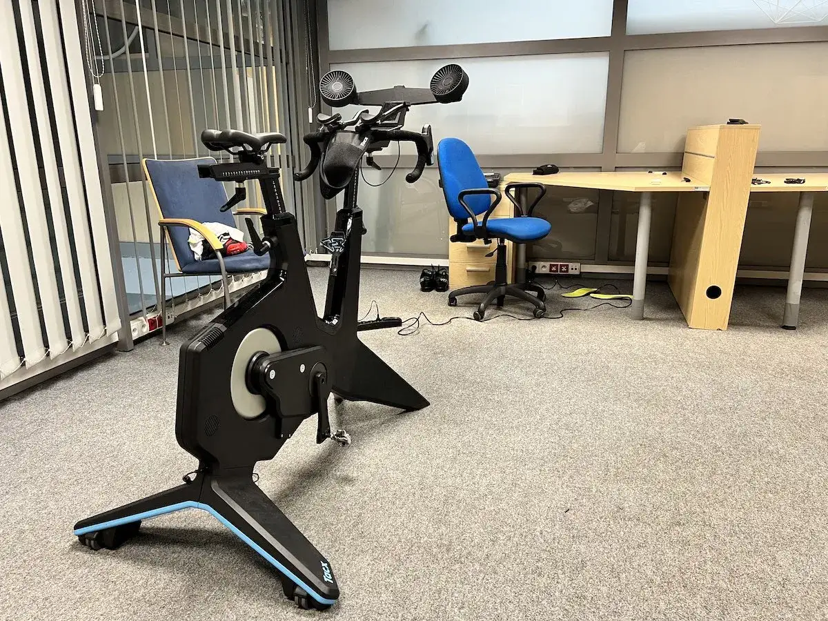 Nowoczesny trenażer rowerowy Tacx, idealny do trening&oacute;w FTP w domu. Obok biurko i krzesła biurowe.