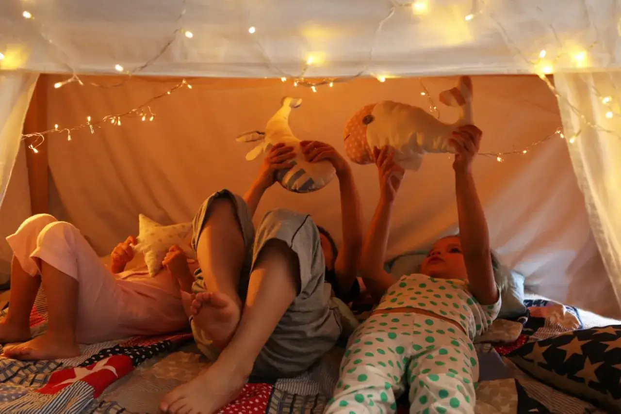 Dzieci w przytulnej bazie z kocy i poduszek, bawią się pluszowymi wielorybami pod migoczącymi światełkami.