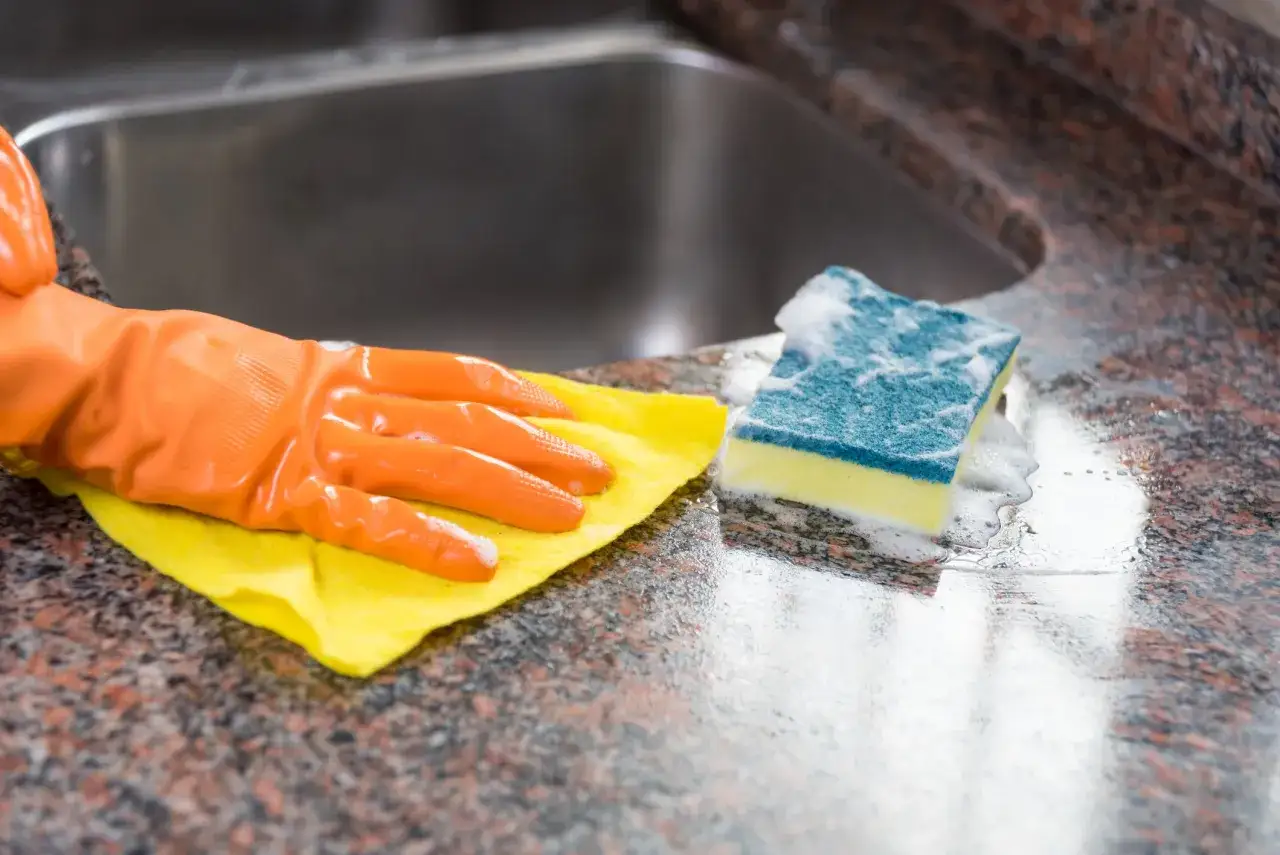 Ręka w pomarańczowej rękawiczce czyści granitową blat octem. Obok gąbka z pianą.