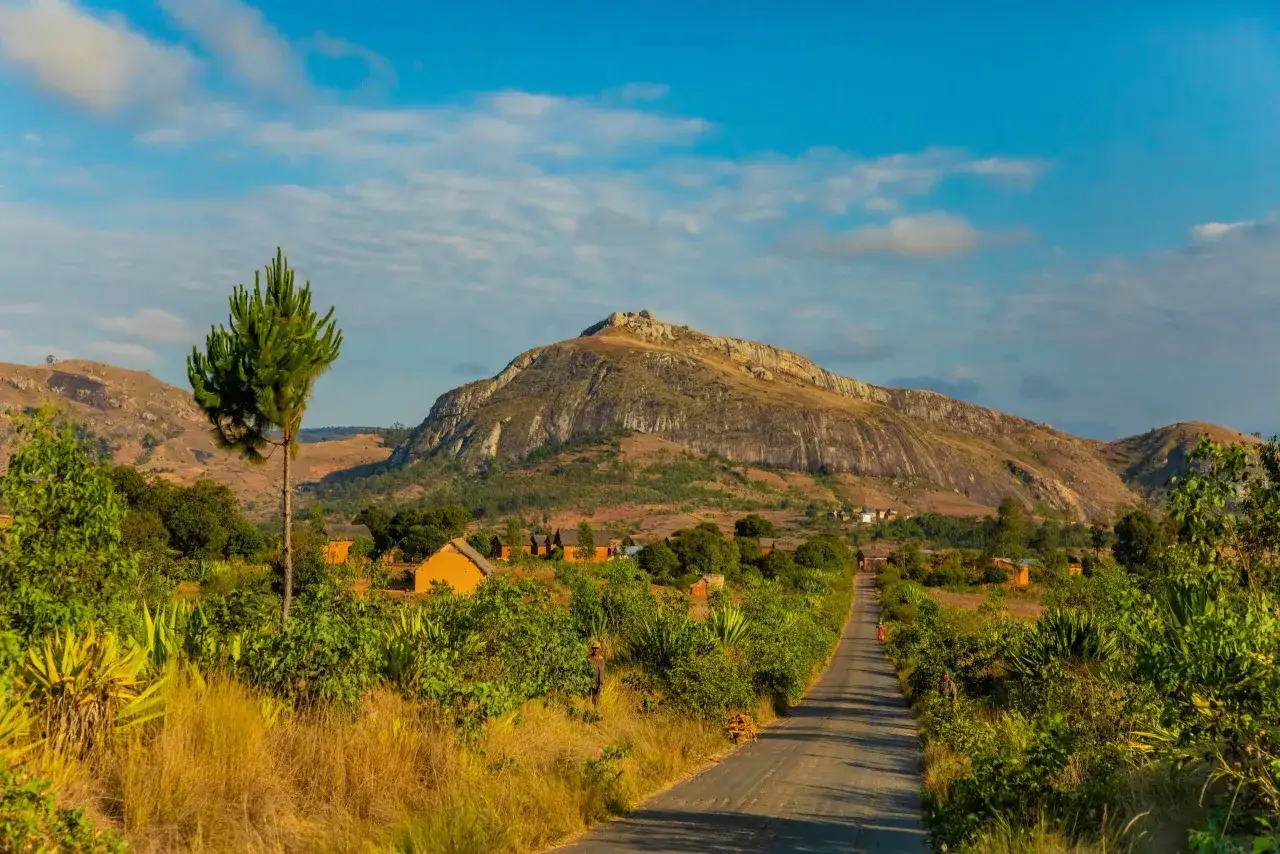 Droga prowadzi do wioski u podn&oacute;ża g&oacute;ry. Zielona roślinność i tradycyjne domy tworzą malowniczy krajobraz, pokazując jedne z wielu **Madagaskar atrakcje**.