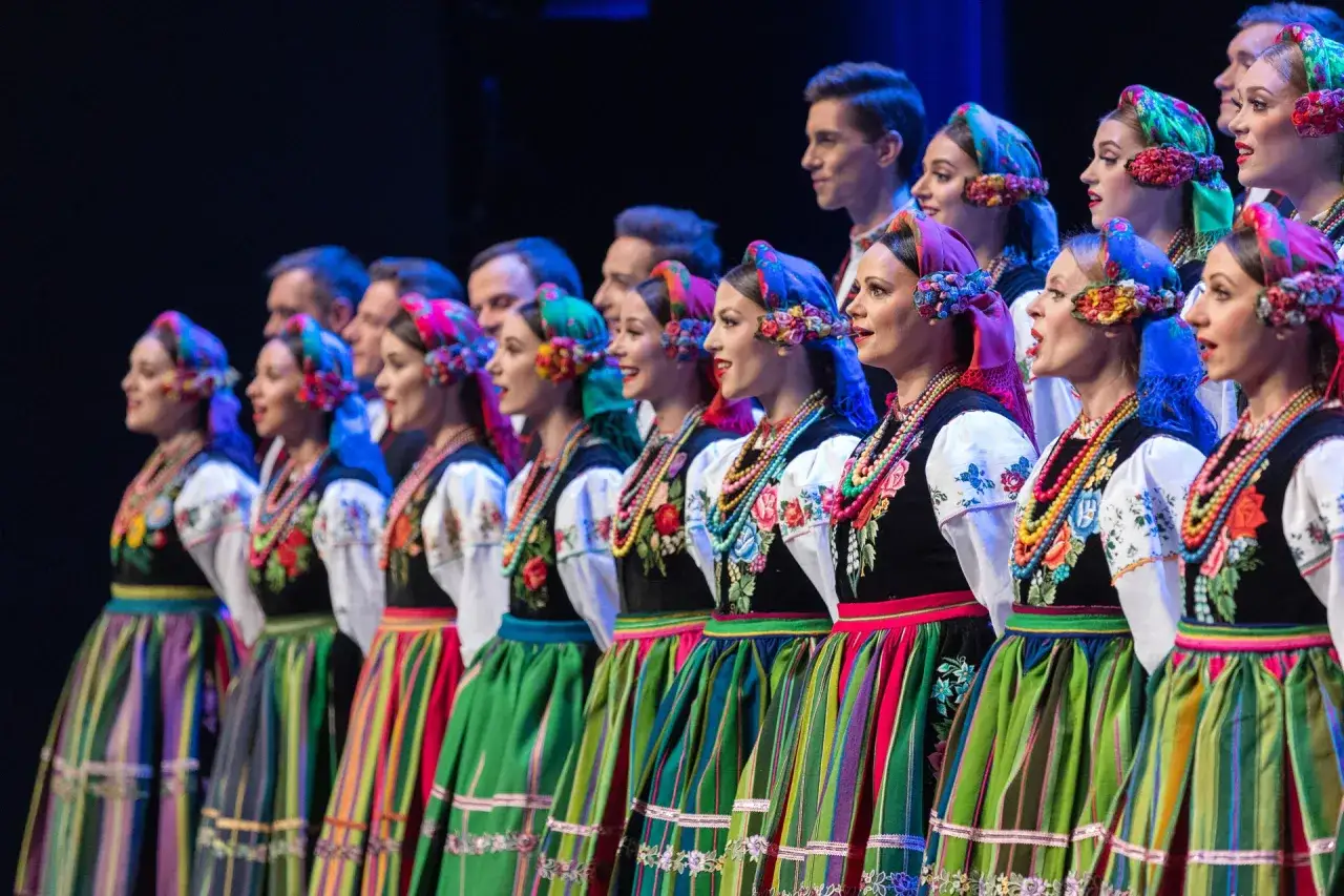 Zespół prezentuje muzykę ludową Mazowsza w barwnych strojach. Kobiety w tradycyjnych chustach i haftowanych gorsetach, mężczyźni w tle.