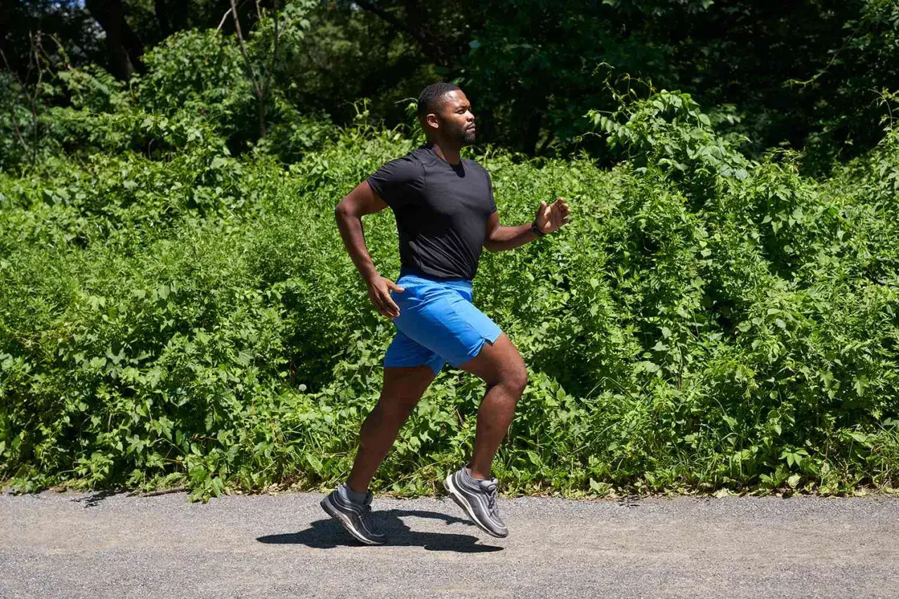 Mężczyzna w niebieskich spodenkach i czarnej koszulce doskonali technikę biegu na ścieżce wśród zieleni.