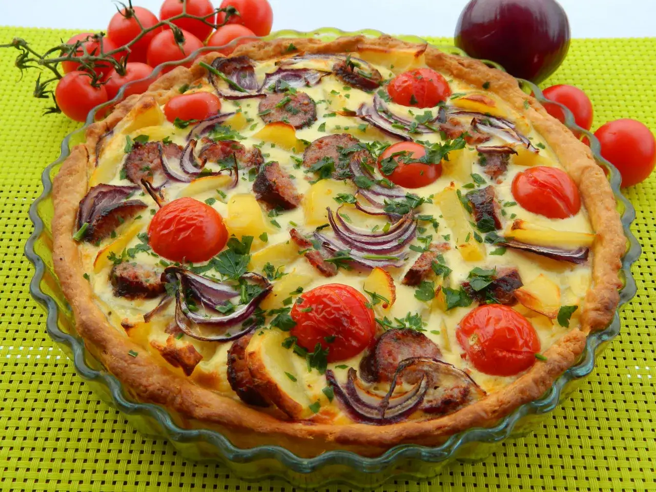 Pyszna tarta z kiełbasą, ziemniakami, pomidorkami i czerwoną cebulą, posypana natką pietruszki.