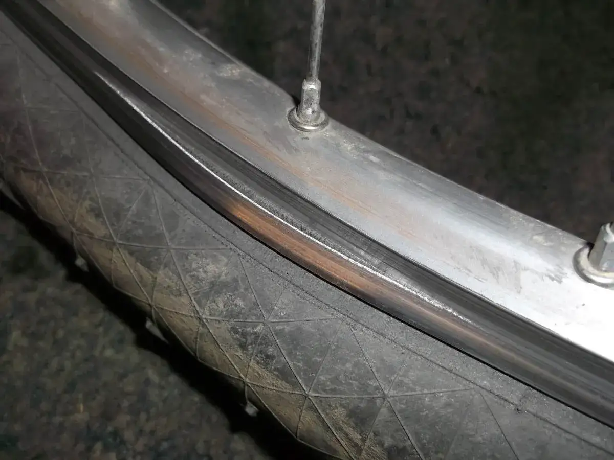 Widok z bliska na zgiętą obręcz koła rowerowego, kt&oacute;ra wymaga prostowania. Widać zużytą oponę i metalową felgę.