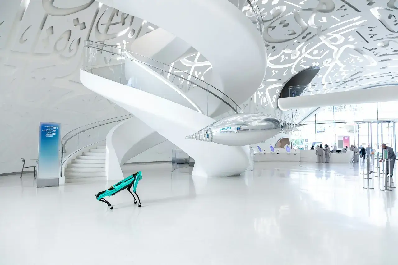 Futurystyczny robot przemierza białe wnętrze Muzeum Przyszłości w Dubaju, podwieszony srebrny obiekt Festo i ludzie w tle.