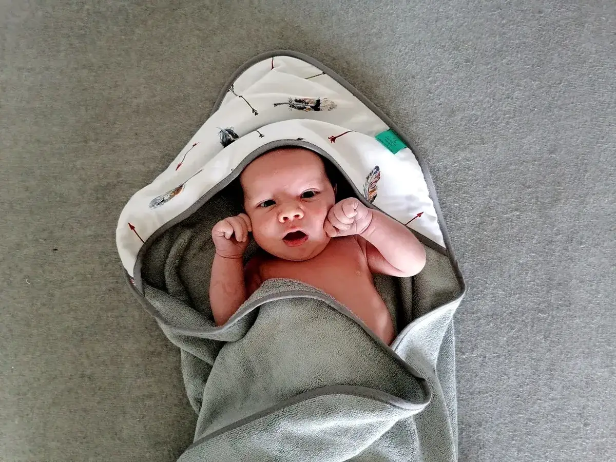 Uroczy niemowlak otulony w miękki, szary ręcznik kąpielowy dla niemowląt z kapturem ozdobionym wzorem strzałek i piór.