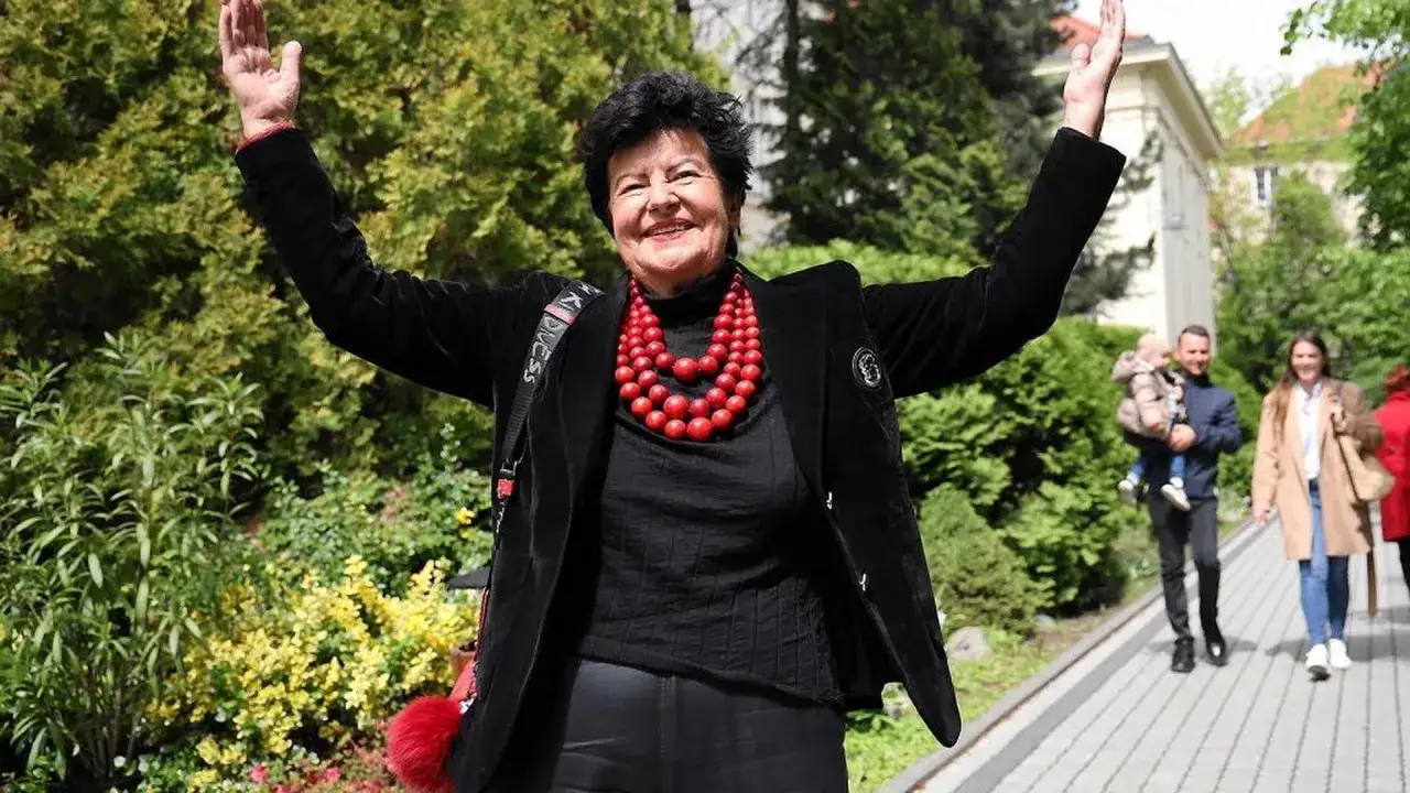 Joanna Senyszyn, mimo że chora, radośnie wita ludzi, machając rękami. Uśmiechnięta, w czarnym stroju i czerwonej koralowej naszyjniku.