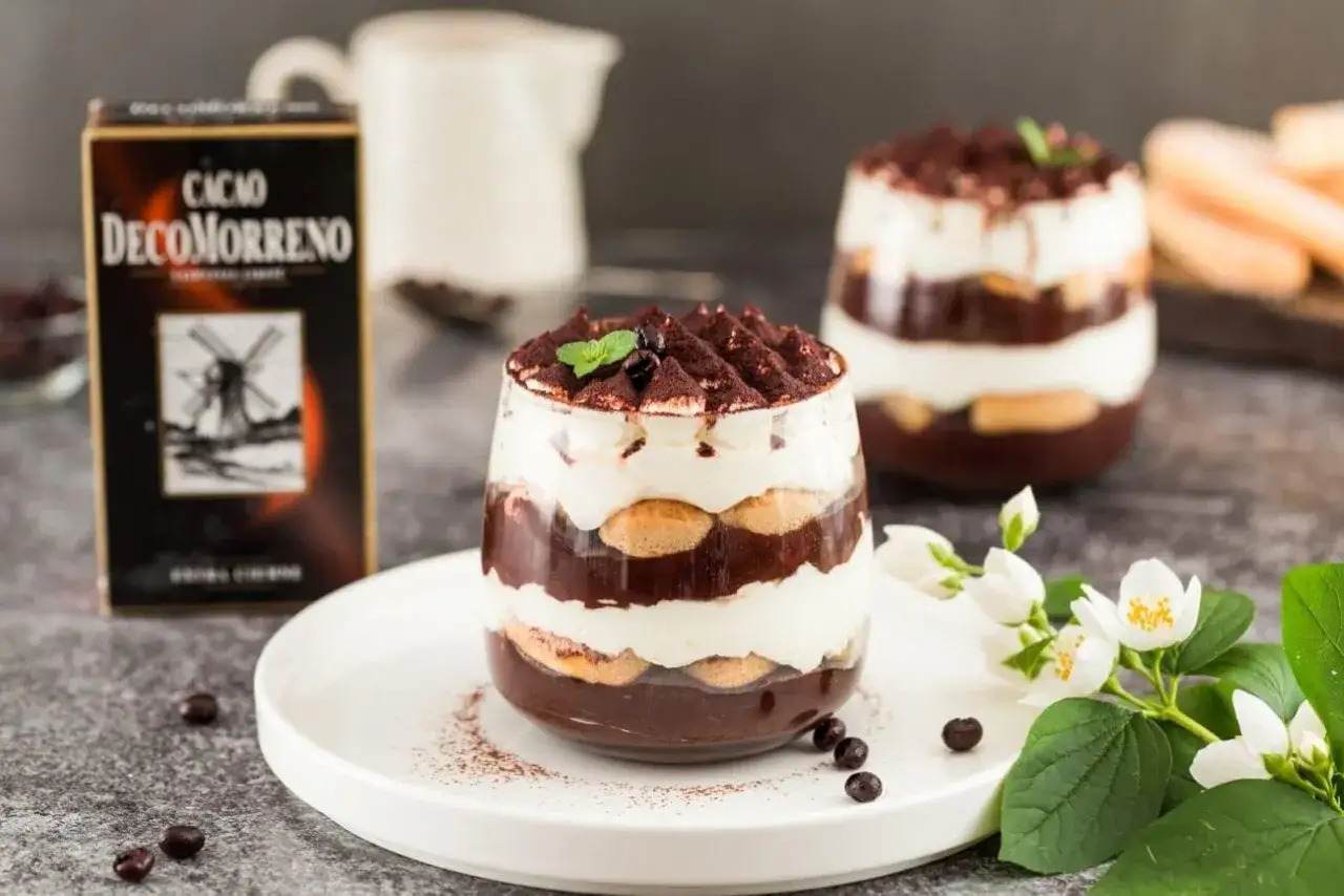 Pyszne tiramisu czekoladowe w szklance, warstwowo ułożone z kremem i biszkoptami, posypane kakao. Obok pudełko Cacao DecoMorreno.