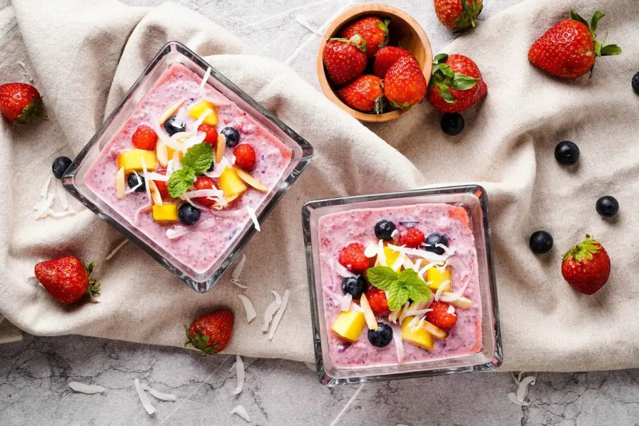 Dwa pucharki z puddingiem chia na słodko, udekorowane owocami leśnymi, mango i wiórkami kokosowymi.
