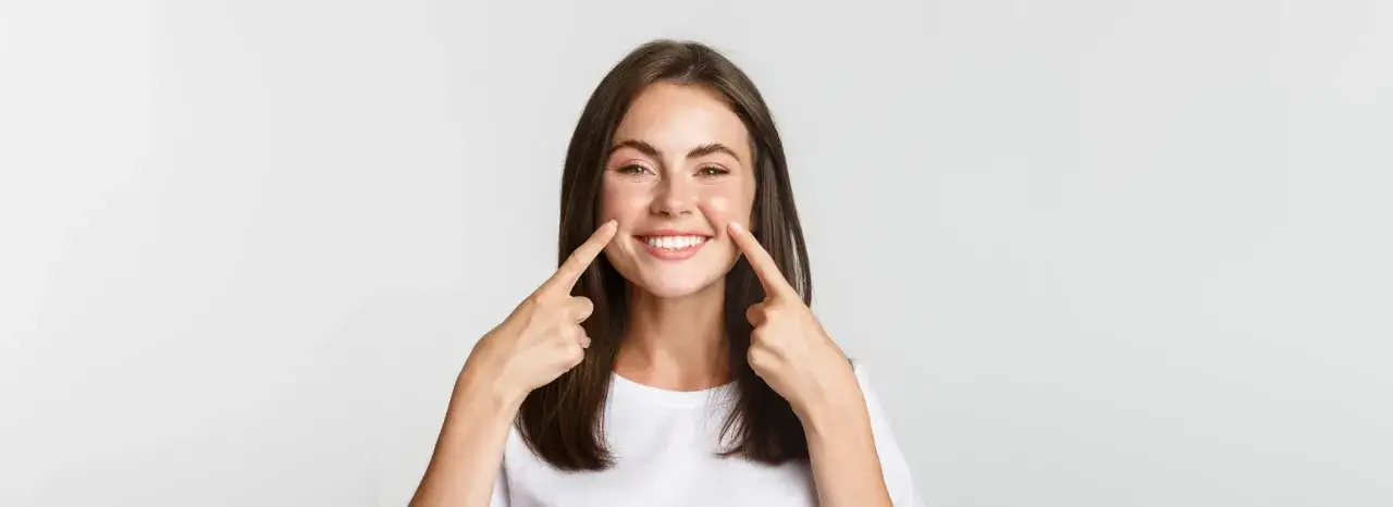 Uśmiechnięta dziewczyna z zalotnym uśmiechem wskazuje palcami na swoje policzki, podkreślając swoją radość i pewność siebie.