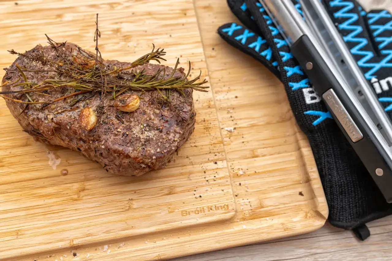 Soczysty stek z rostbefu, doprawiony ziołami i czosnkiem, leży na desce obok rękawic grillowych.