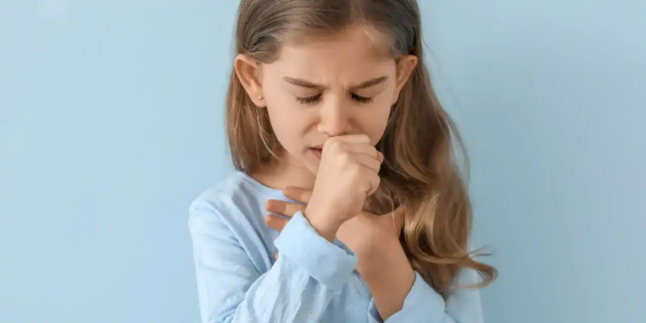 Dziewczynka z zamkniętymi oczami kaszle, trzymając dłoń przy ustach i klatce piersiowej. To objaw mokrego kaszlu u dziecka.