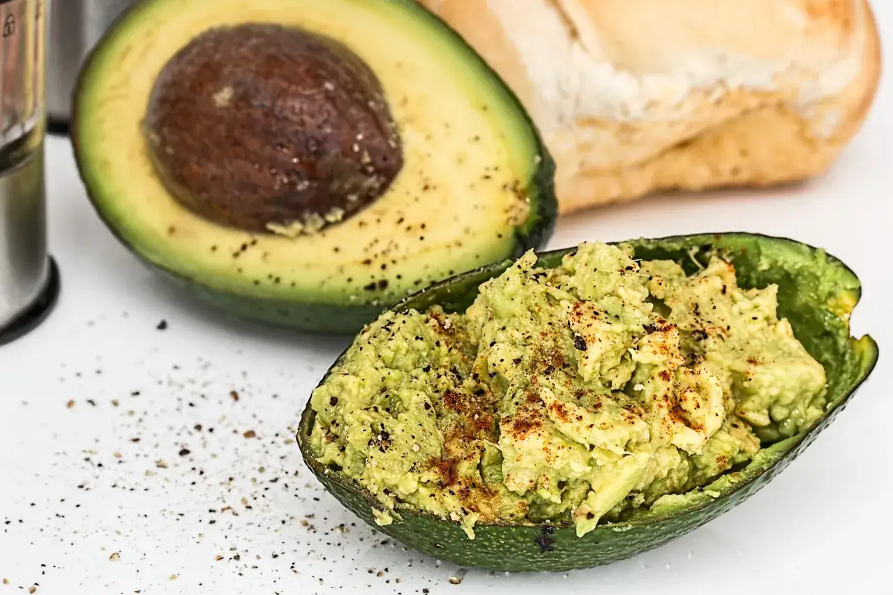 Idealne awokado do guacamole: poł&oacute;wka z pestką i gotowe guacamole w sk&oacute;rce, posypane pieprzem. Obok bułka.
