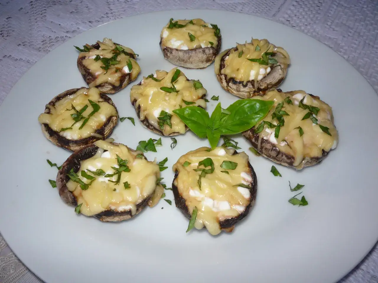 Osiem pieczarek faszerowanych na grilla, zapieczonych z serem i posypanych ziołami, prezentuje się apetycznie na białym talerzu.