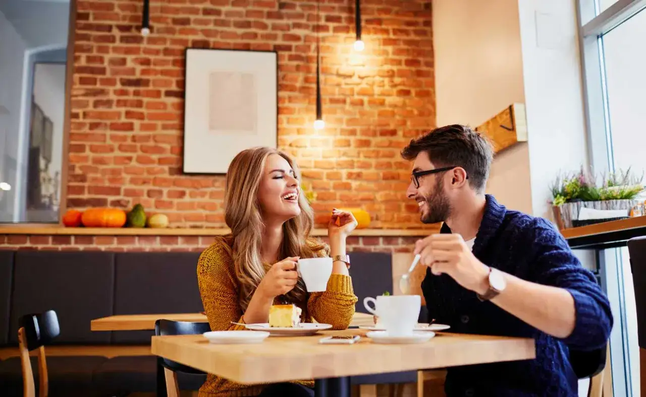 Uśmiechnięta blondynka i mężczyzna przy kawie. Widać, jak facet zaczepia kobietę, a ona śmieje się wesoło.