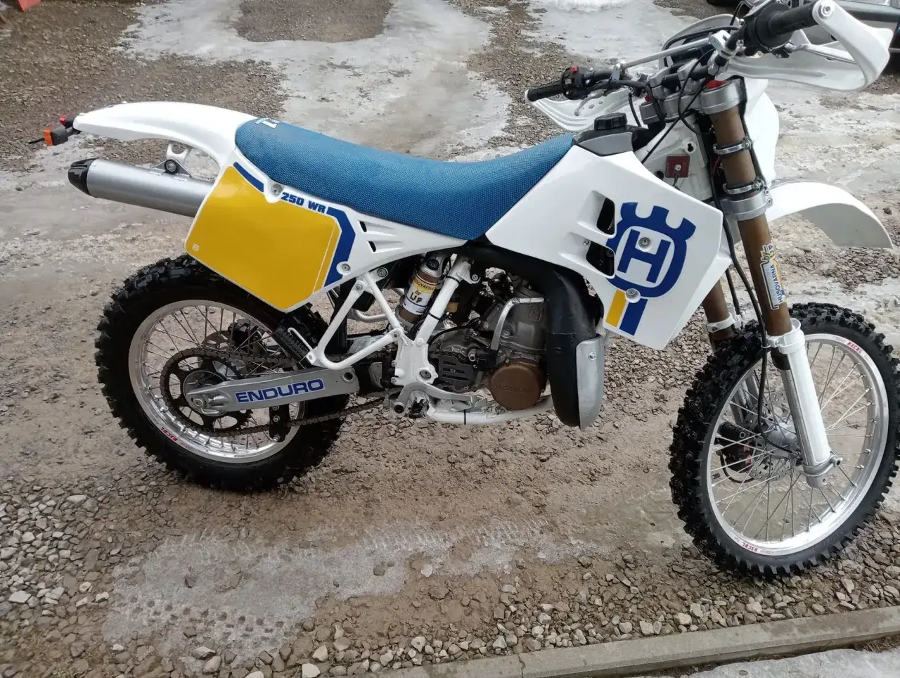 Klasyczny Husqvarna CR 250 Enduro gotowy na przygodę. Biało-ż&oacute;łta grafika i niebieskie siedzenie.