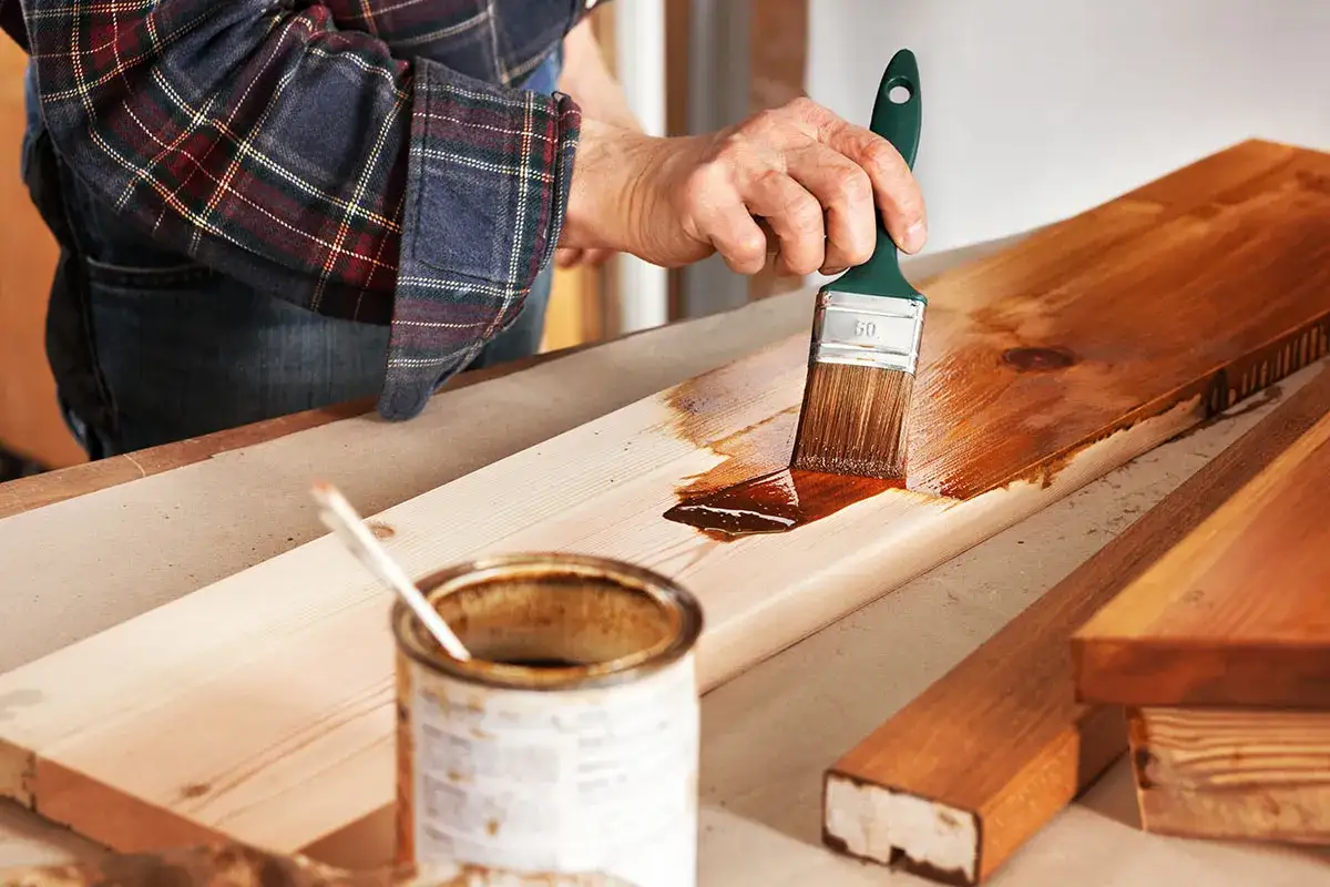 Nakładanie lakieru na schody drewniane. Pędzel rozprowadza brązowy lakier po desce, nadając jej piękny odcień.