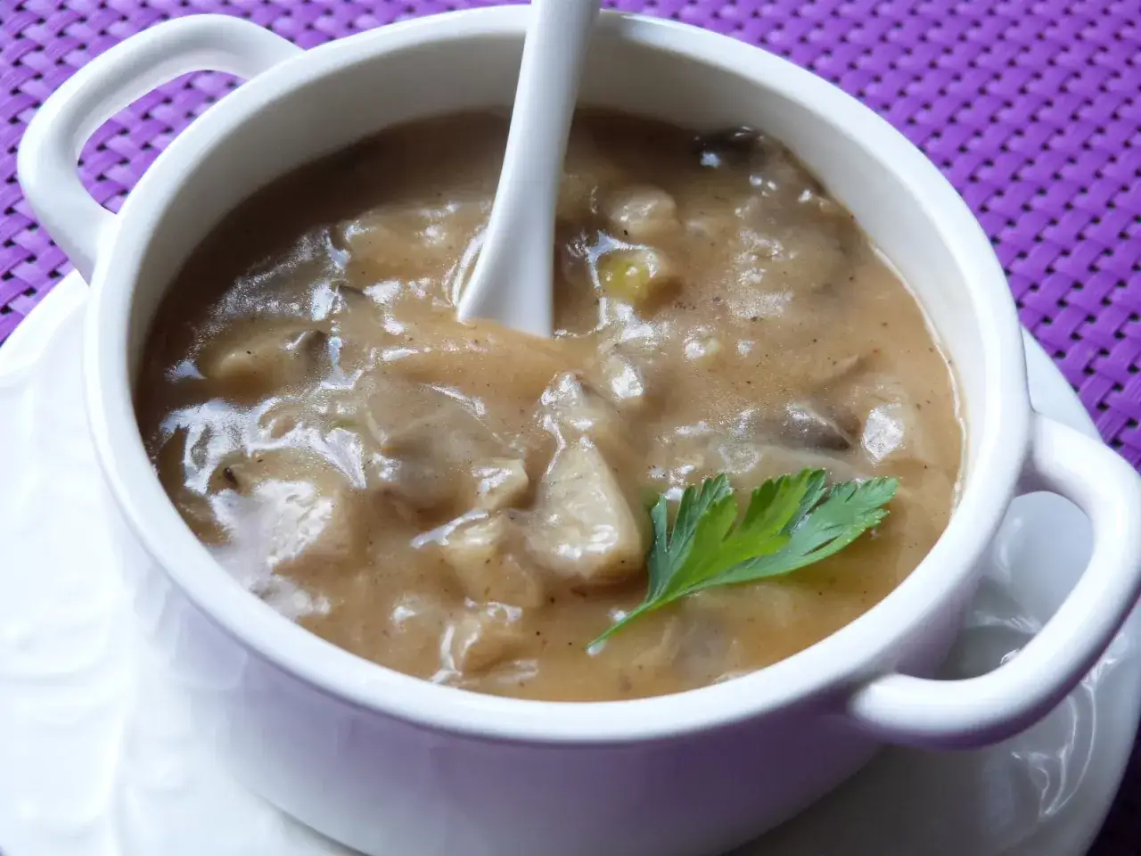 Gęsty, kremowy sos z boczniak&oacute;w, udekorowany listkiem pietruszki, czeka w białej miseczce na łyżkę.