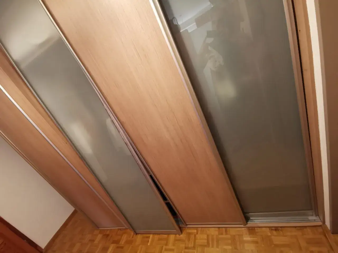 Widok na drzwi przesuwne w szafie, z widoczną regulacją ich położenia. Drewniane fronty i matowe szkło.