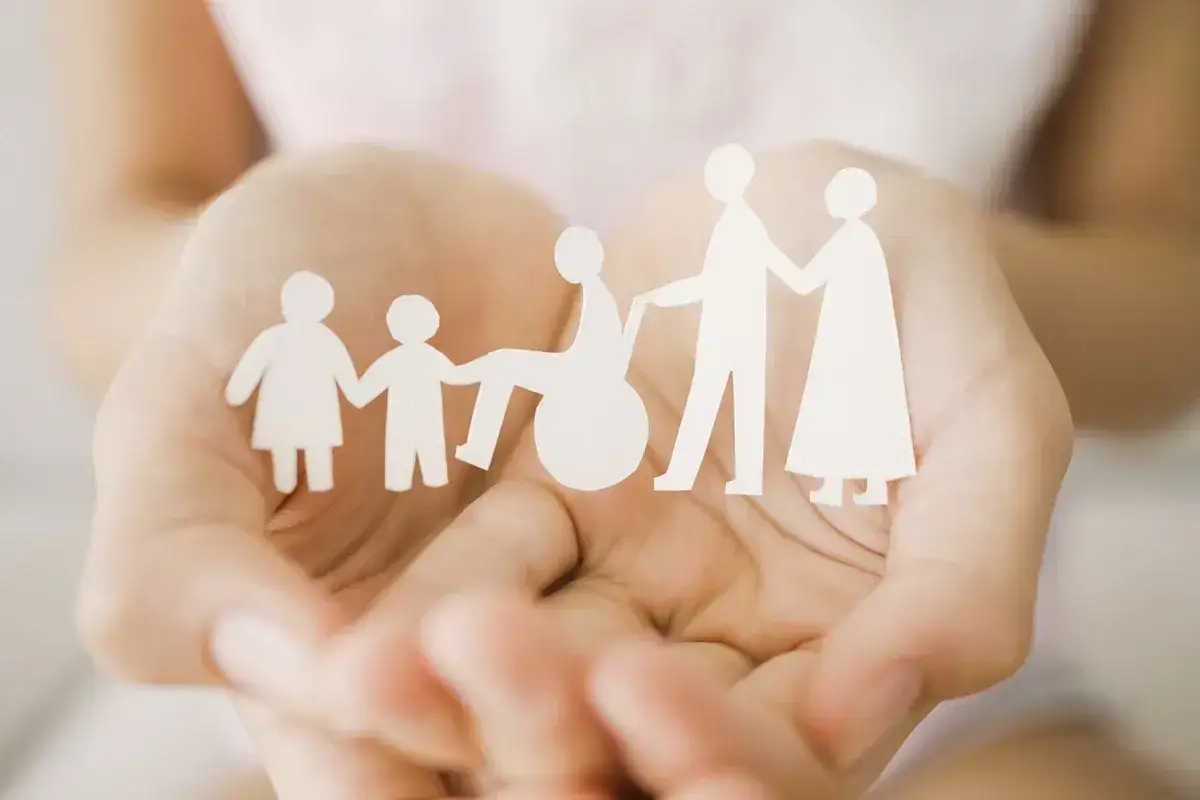 Dłonie trzymają wycięte z papieru postacie symbolizujące rodzinę, w tym osobę na wózku. To obraz wsparcia, jak co to jest zasiłek rodzinny.