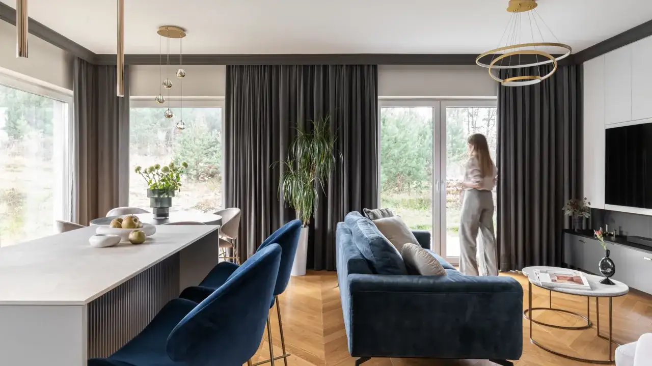 Nowoczesny, elegancki dom z widokiem na las. Granatowa sofa, wyspa kuchenna i stylowe oświetlenie tworzą przytulną atmosferę.