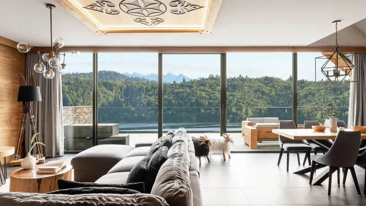 Luksusowy salon z widokiem na g&oacute;ry i jezioro. Dwie owieczki stoją obok kanapy. To jest właśnie glamping &ndash; komfort w otoczeniu natury.