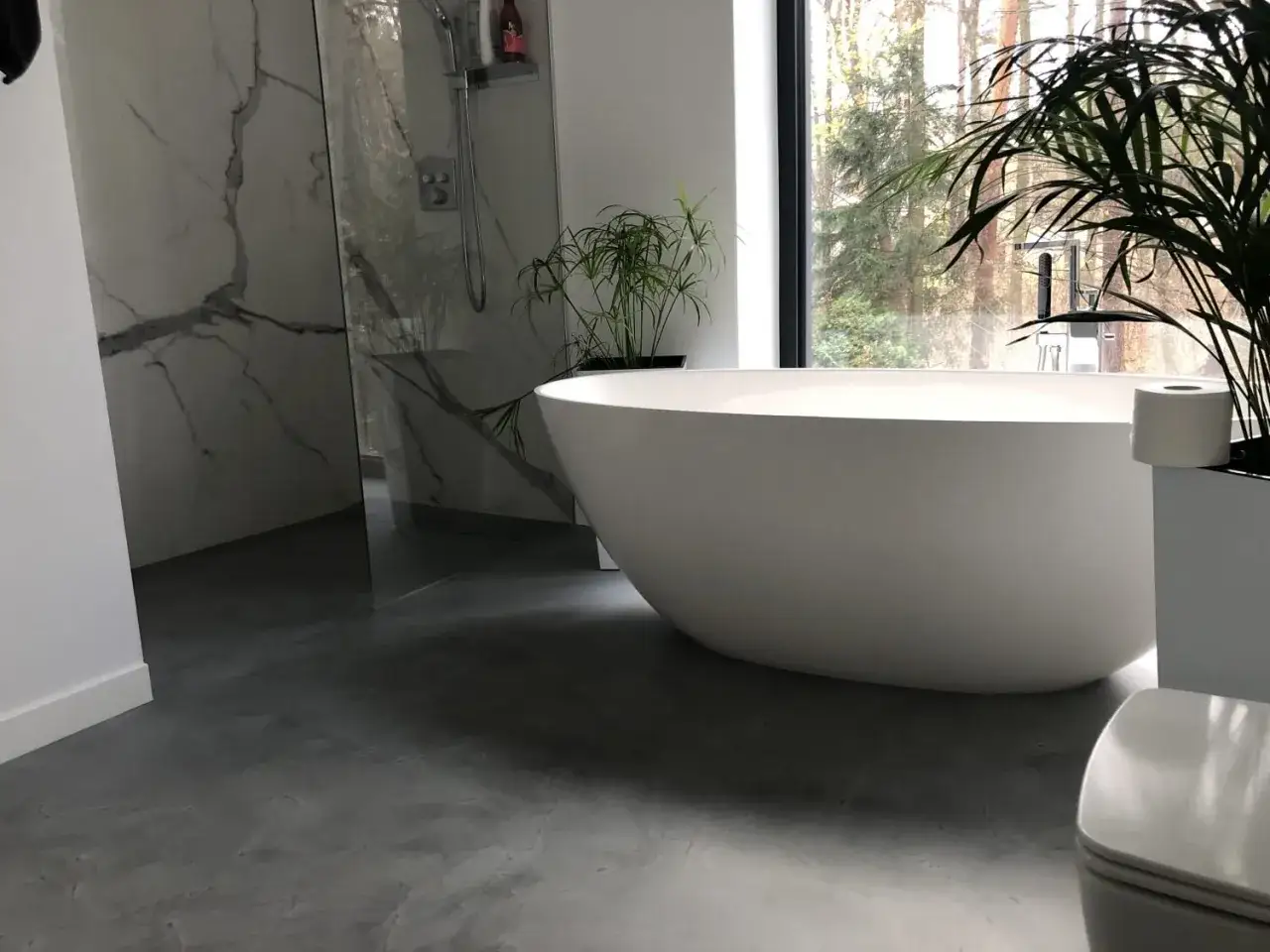 Nowoczesna łazienka z wanną wolnostojącą i prysznicem. Podłoga wykonana z materiału przypominającego beton, czyli mikrocementu, nadaje wnętrzu surowego charakteru.