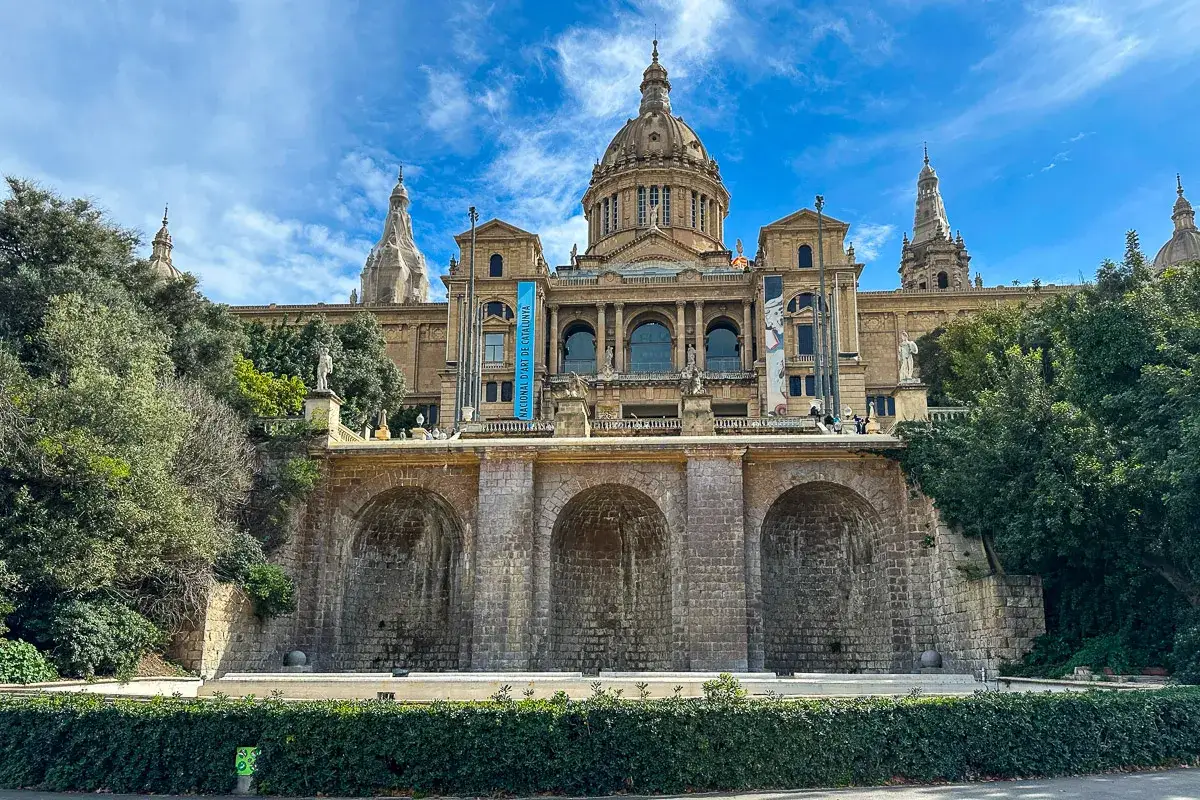 Majestatyczny budynek muzeum sztuki w Barcelonie, z kopułą i arkadami, otoczony zielenią.