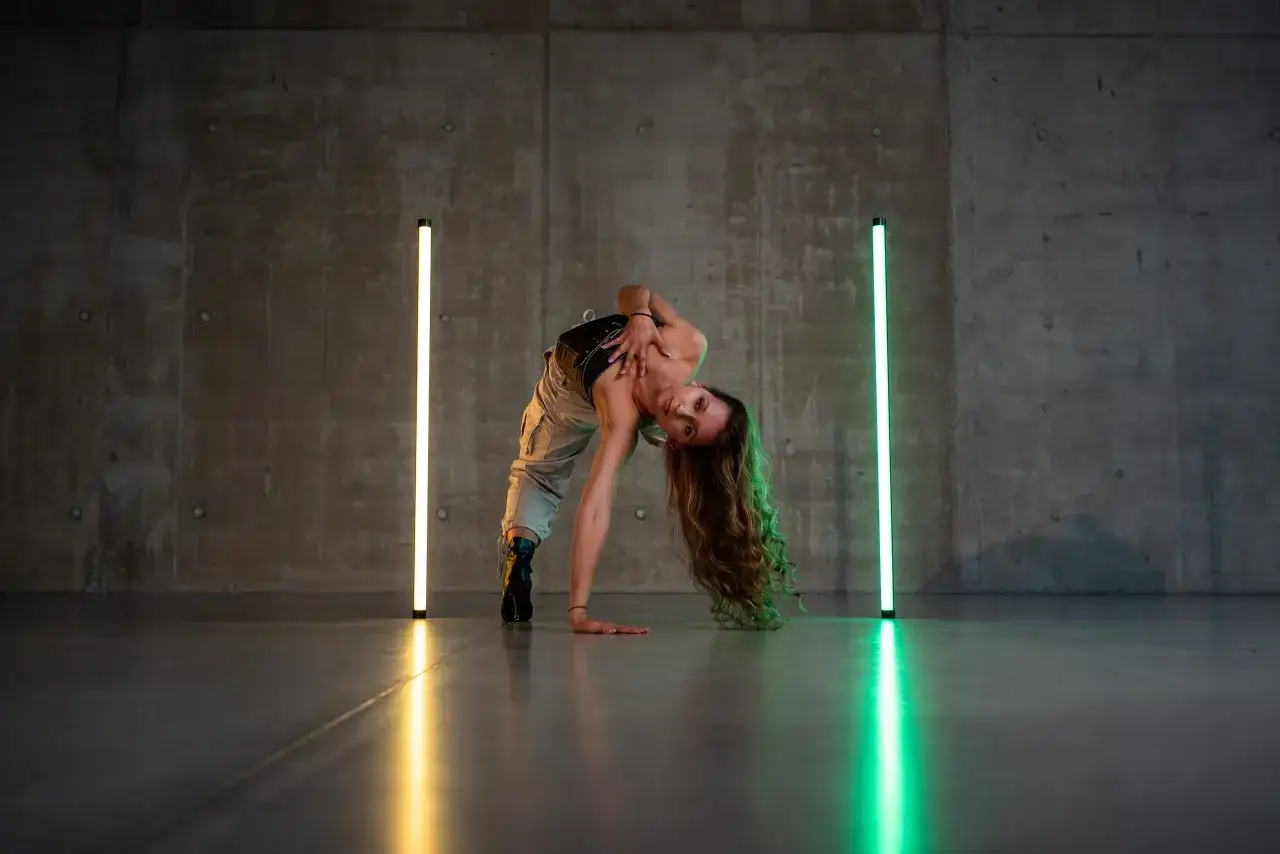 Tancerka w pozie łuku, ćwicząca naukę tańca nowoczesnego. Długie włosy opadają na podłogę, a światła neonowe podkreślają dynamikę ruchu.