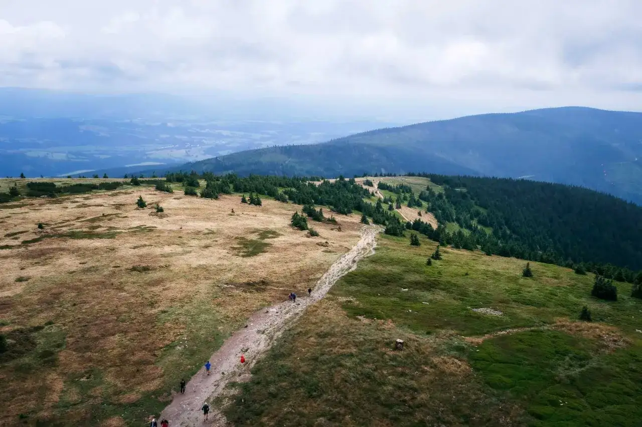 Grupa turyst&oacute;w wędruje kamienistym szlakiem przez malownicze **Beskidy Kłodzko**. W tle rozciągają się zielone wzg&oacute;rza i mgliste doliny.