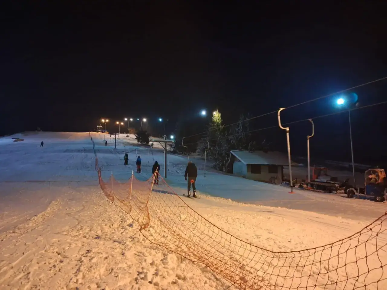 Oświetlony stok narciarski nocą, idealne miejsce, gdzie na narty w Kujawsko-Pomorskiem. Widać narciarzy, wyciąg i ratrak.