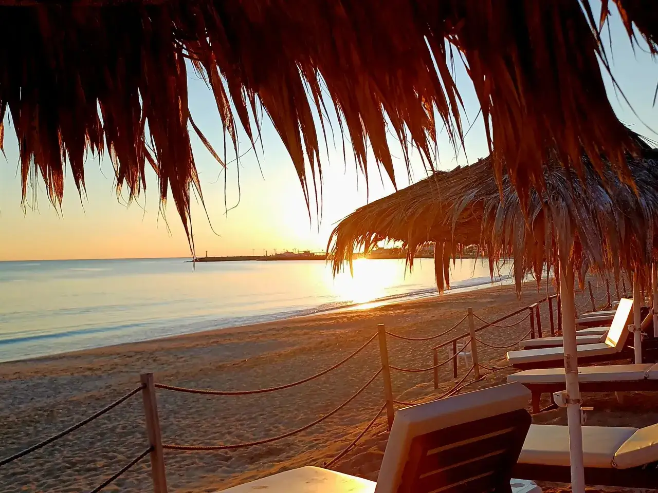 Zach&oacute;d słońca na plaży w Ayia Napa. Leżaki pod słomianymi parasolami, idealne miejsce, by zobaczyć piękno Cypru.