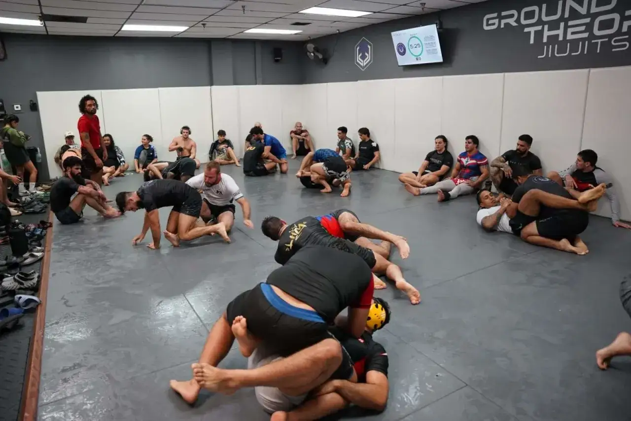 Grupa ćwiczy no gi bjj na macie. Niekt&oacute;rzy siedzą, inni sparują.