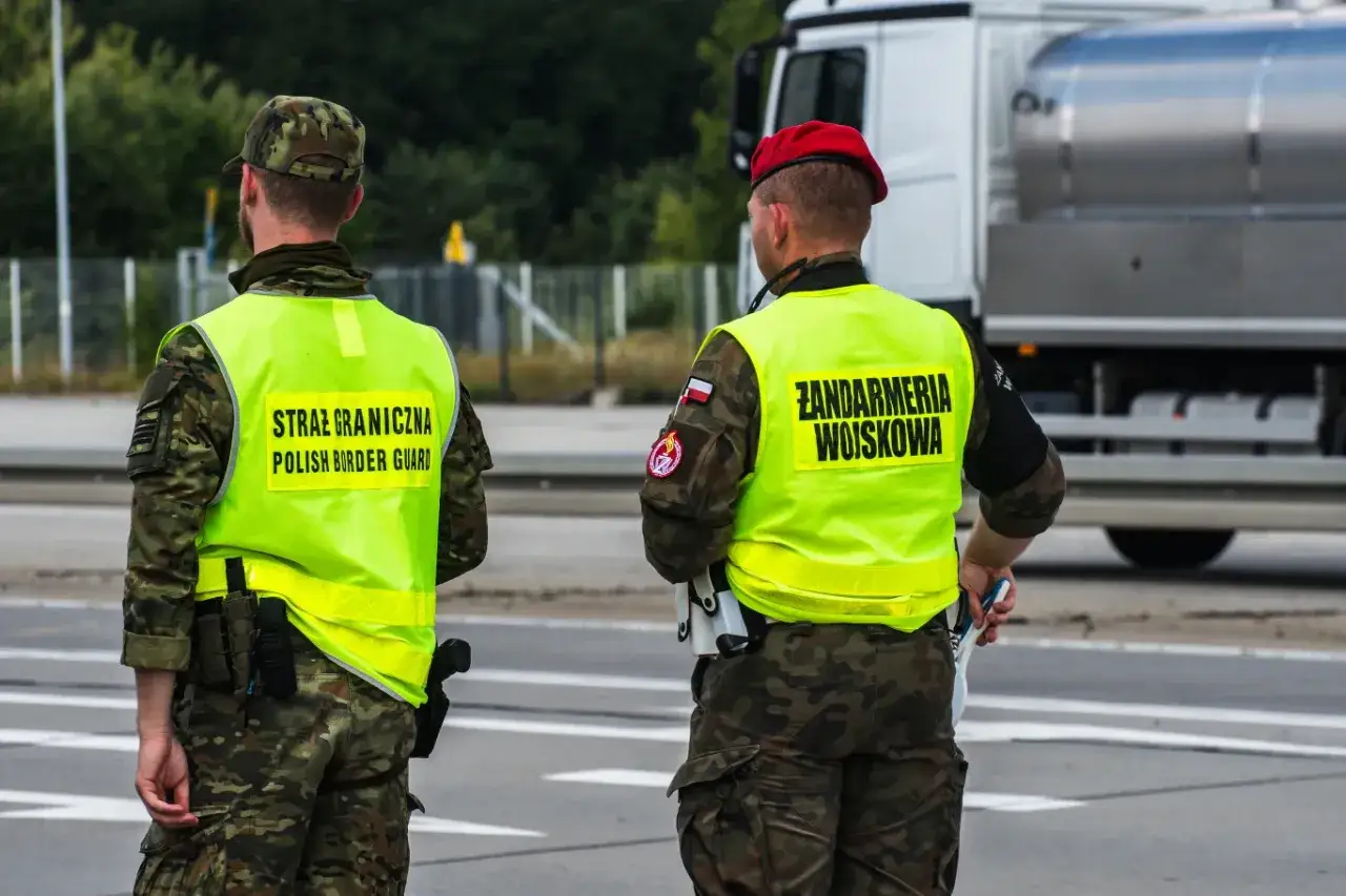 Dwóch funkcjonariuszy służby mundurowej w kamizelkach odblaskowych: Straż Graniczna i Żandarmeria Wojskowa, stoi na drodze.