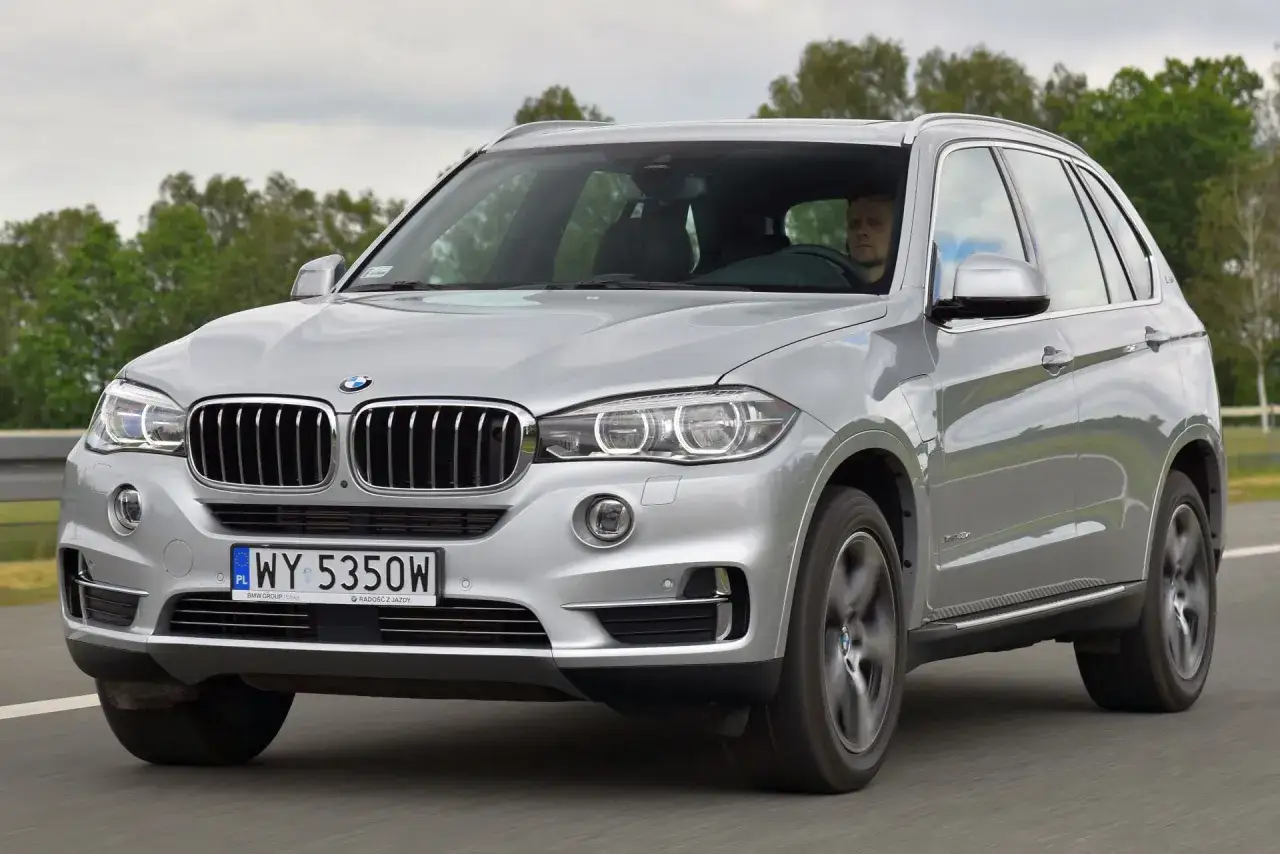 Srebrny BMW X5 na drodze. Kierowca w środku, drzewa w tle. Wiele pozytywnych bmw x 5 opinii potwierdza jego jakość.