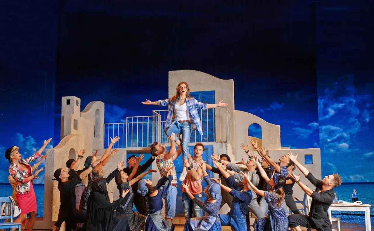 Mamma mia po polsku! Aktorka na scenie, uniesiona przez innych, w otoczeniu chóru. Grecka sceneria i niebieskie niebo.