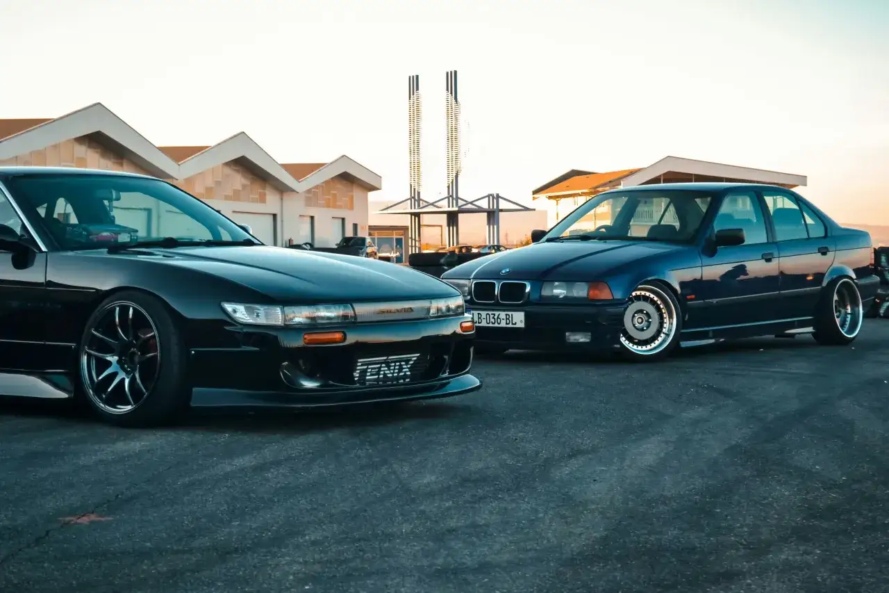 Dwa zmodyfikowane samochody, czarna Nissan Silvia i niebieskie BMW E36, gotowe do akcji. Ich tuning zachwyca.