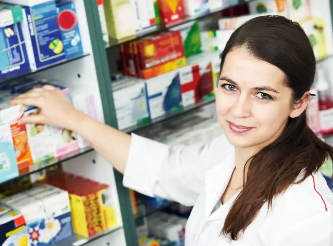 Farmaceutka uśmiecha się, sięgając po lek. Czy leki refundowane wszędzie kosztują tyle samo? Zapytaj w aptece.