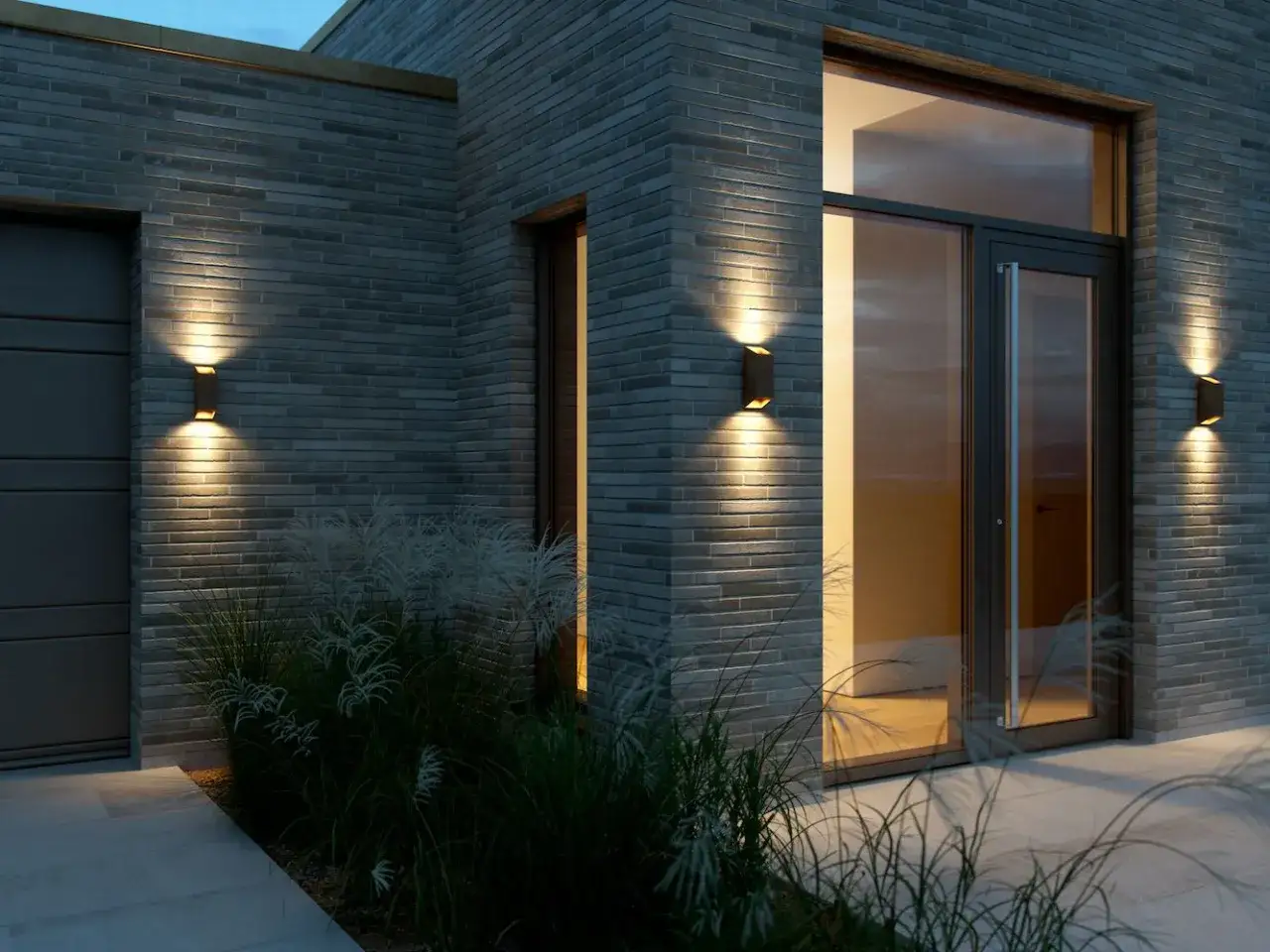 Nowoczesny kinkiet góra dół oświetla szarą ceglaną elewację domu, tworząc ciepłą atmosferę.
