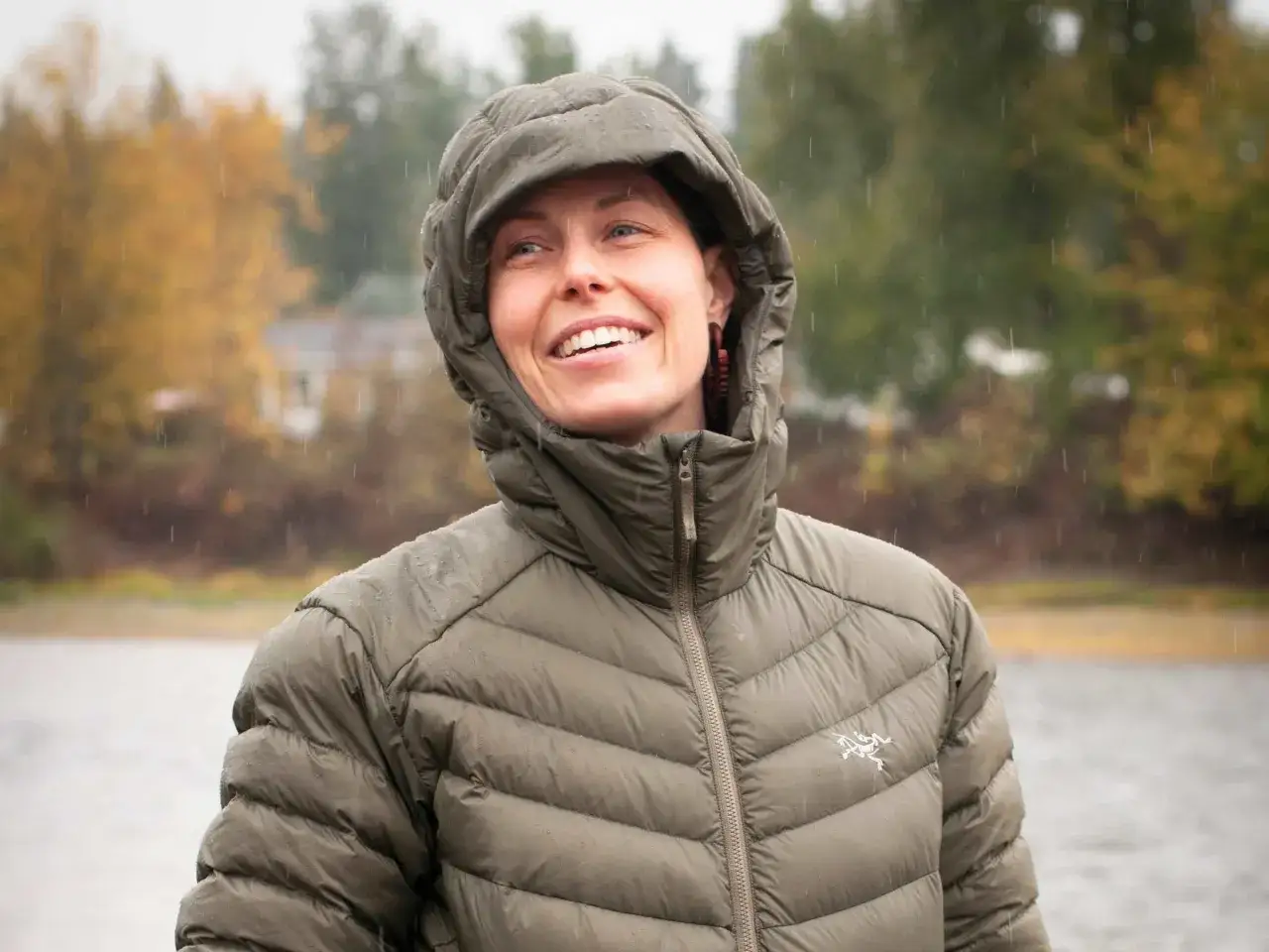 Uśmiechnięta kobieta w kurtce z kapturem, na kt&oacute;rej widać krople deszczu. Kurtka wypełniona jest materiałem Primaloft, kt&oacute;ry zapewnia ciepło nawet w wilgotnych warunkach.