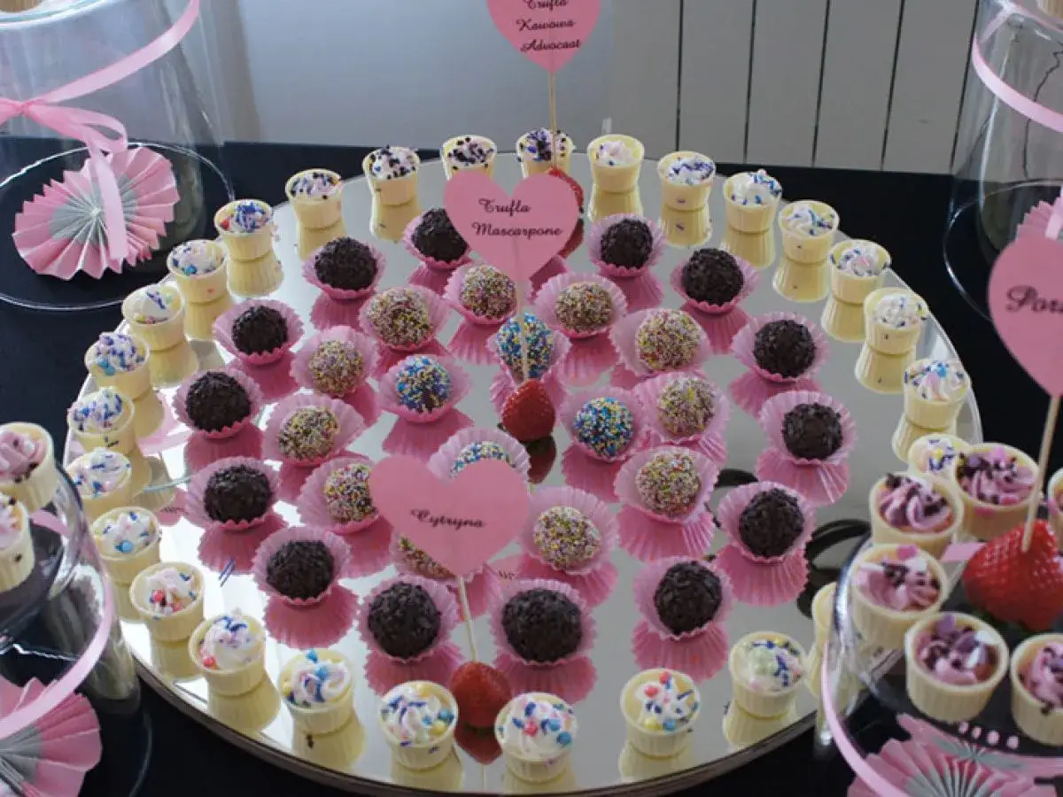 Słodki stół na komunię jak zrobić: kolorowe pralinki i babeczki w różowych papilotkach, ozdobione serduszkami.