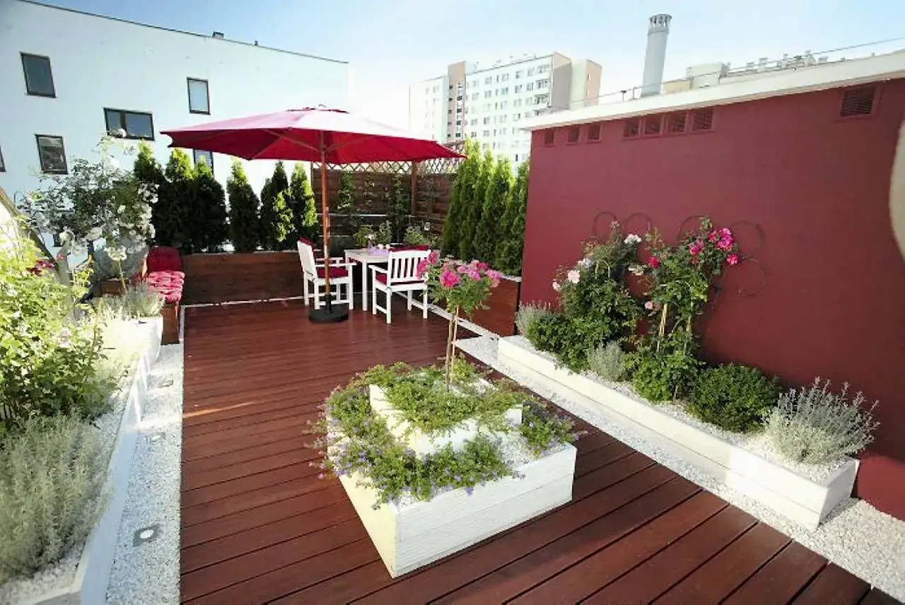 Taras z meblami i parasolem, idealne miejsce na relaks. Wokół dużo zieleni, kwiatów i białych donic. Co taniego na taras? Tutaj jest pięknie i stylowo.