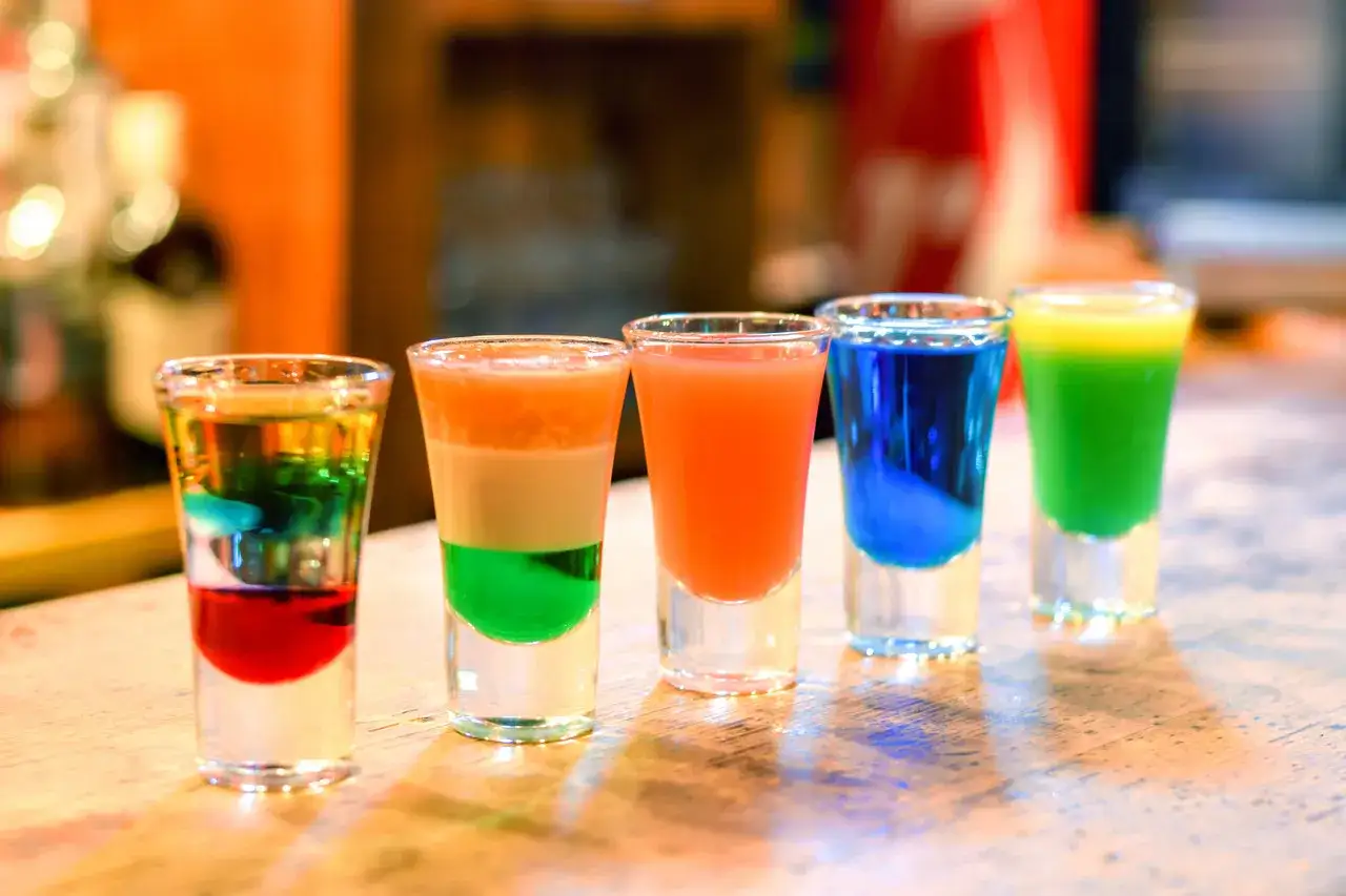 Pięć kolorowych shotów na barze: jeden wielowarstwowy, jeden z trzema warstwami, jeden pomarańczowy, jeden niebieski i jeden zielony. Shoty warstwowe kontra shoty mieszane.