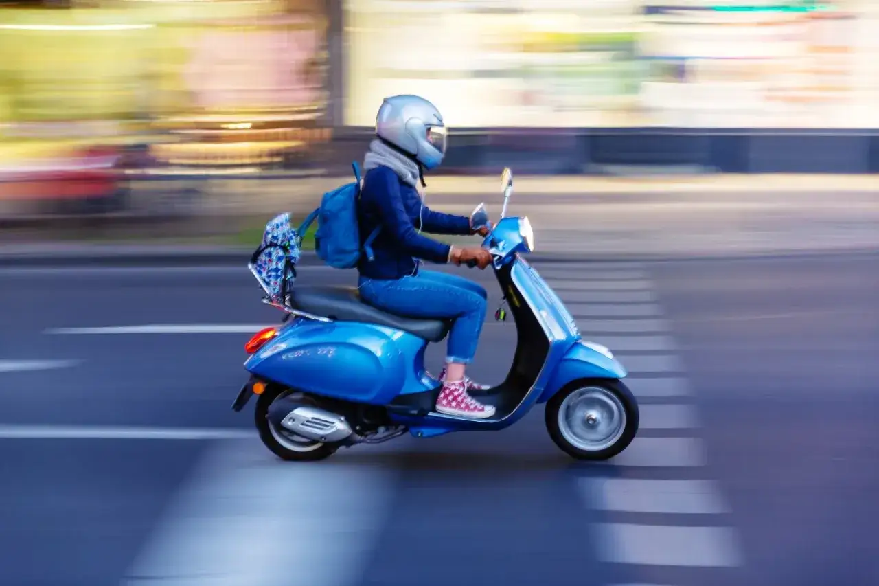 Kobieta w kasku jedzie niebieskim skuterem 125. Czy skuterem 125 można jeździć po autostradzie? To pytanie nurtuje wielu kierowc&oacute;w.